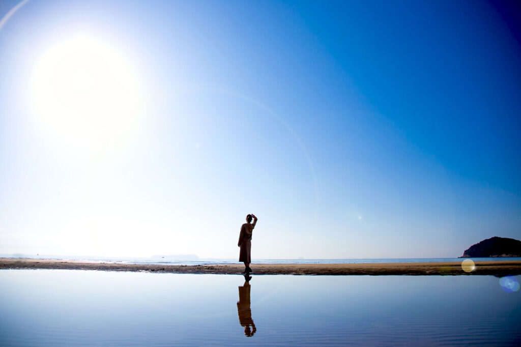 青空の下、鏡のように静かな水面に人影が反射している海岸の風景。太陽の光が強く差し込み、人物は逆光でシルエットのように写っている。